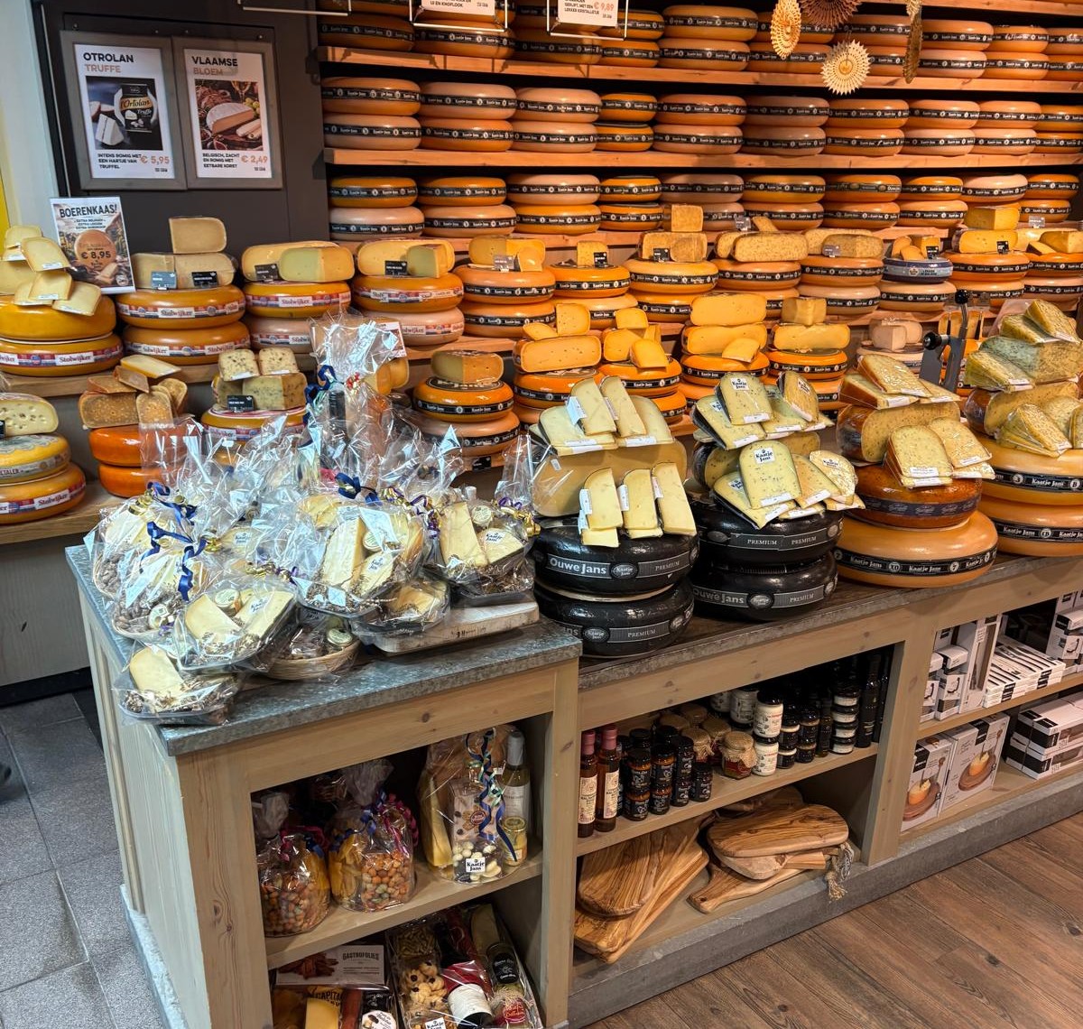 Houten stellingen en een toonbank volledig gevuld met diverse soorten ronde kazen, verpakte kaasstukjes en geschenkpakketten.