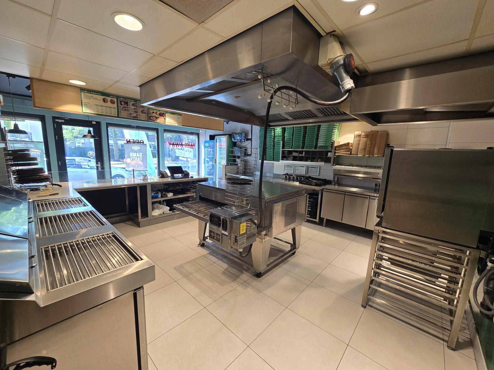 Professionele keuken met roestvrijstalen werkbanken, een grote pizzaoven onder een afzuigkap en opgestapelde groene pizzadozen op de achtergrond.