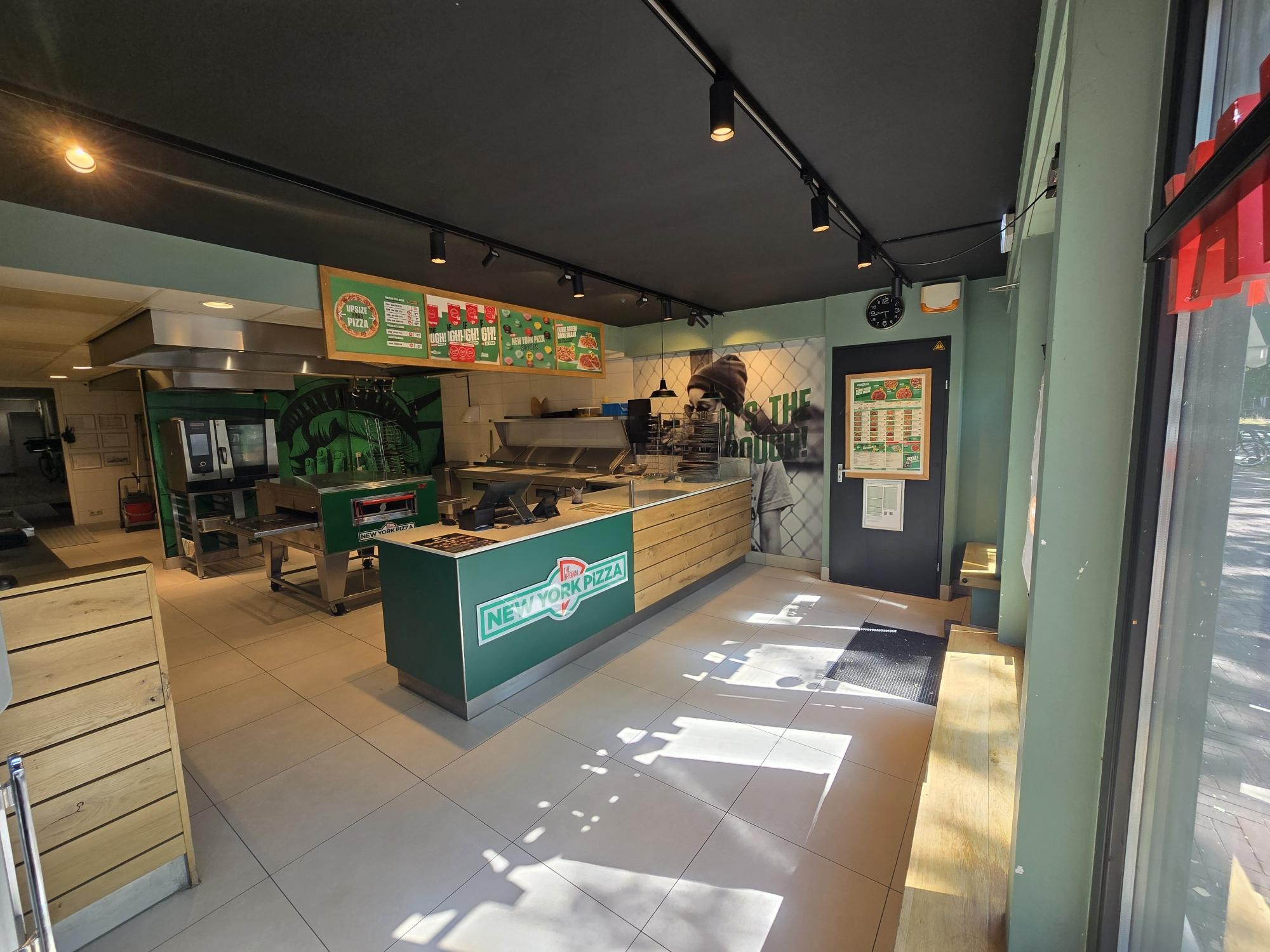 Interieur van de winkel met een groene New York Pizza balie, een menukaart boven de werkplek en een tegelvloer verlicht door zonlicht.