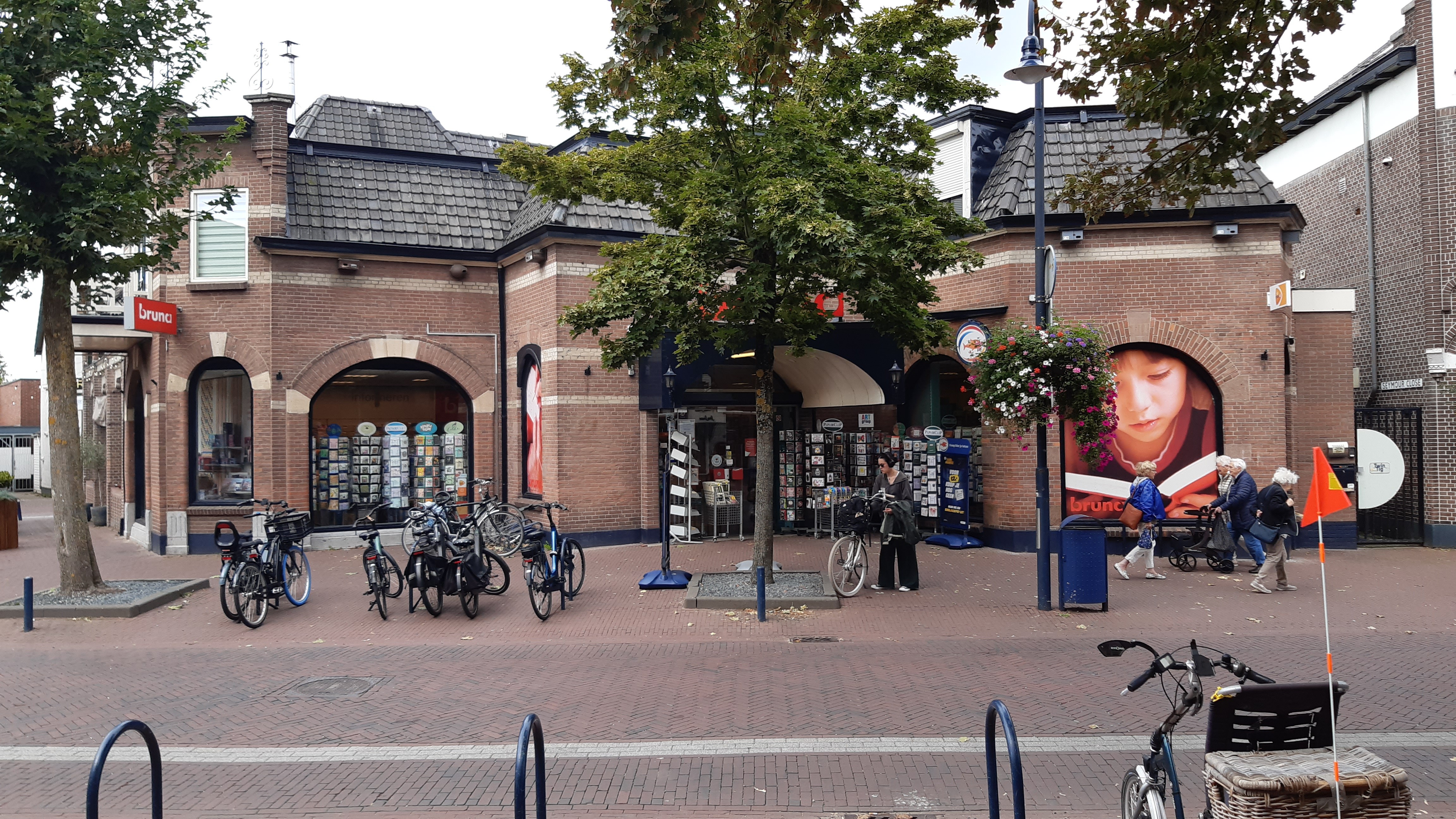 Vooraanzicht van het bakstenen winkelpand van Bruna met grote etalageramen, geparkeerde fietsen en voorbijlopende mensen.