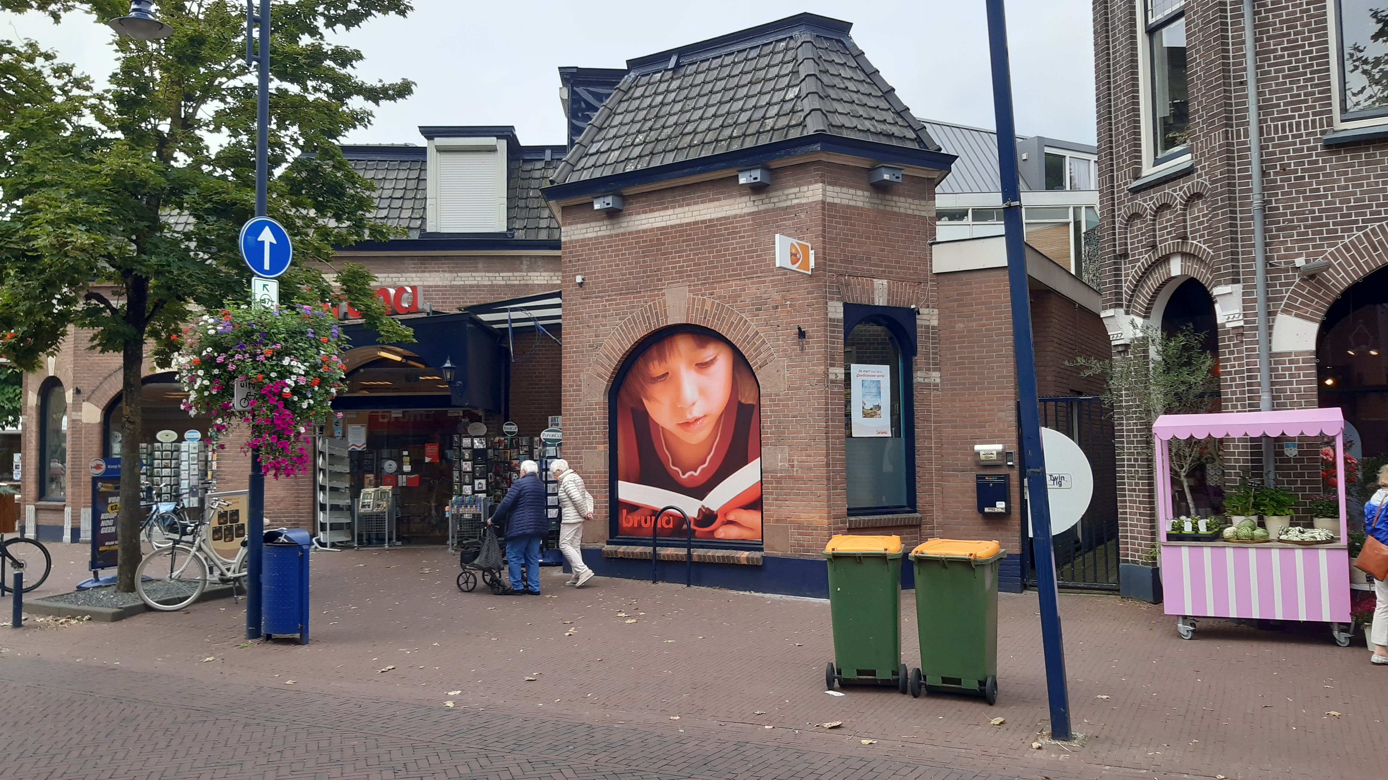 Hoek van het bakstenen pand met een grote raamafbeelding van een lezend kind, twee groene afvalcontainers en een roze marktkraam.