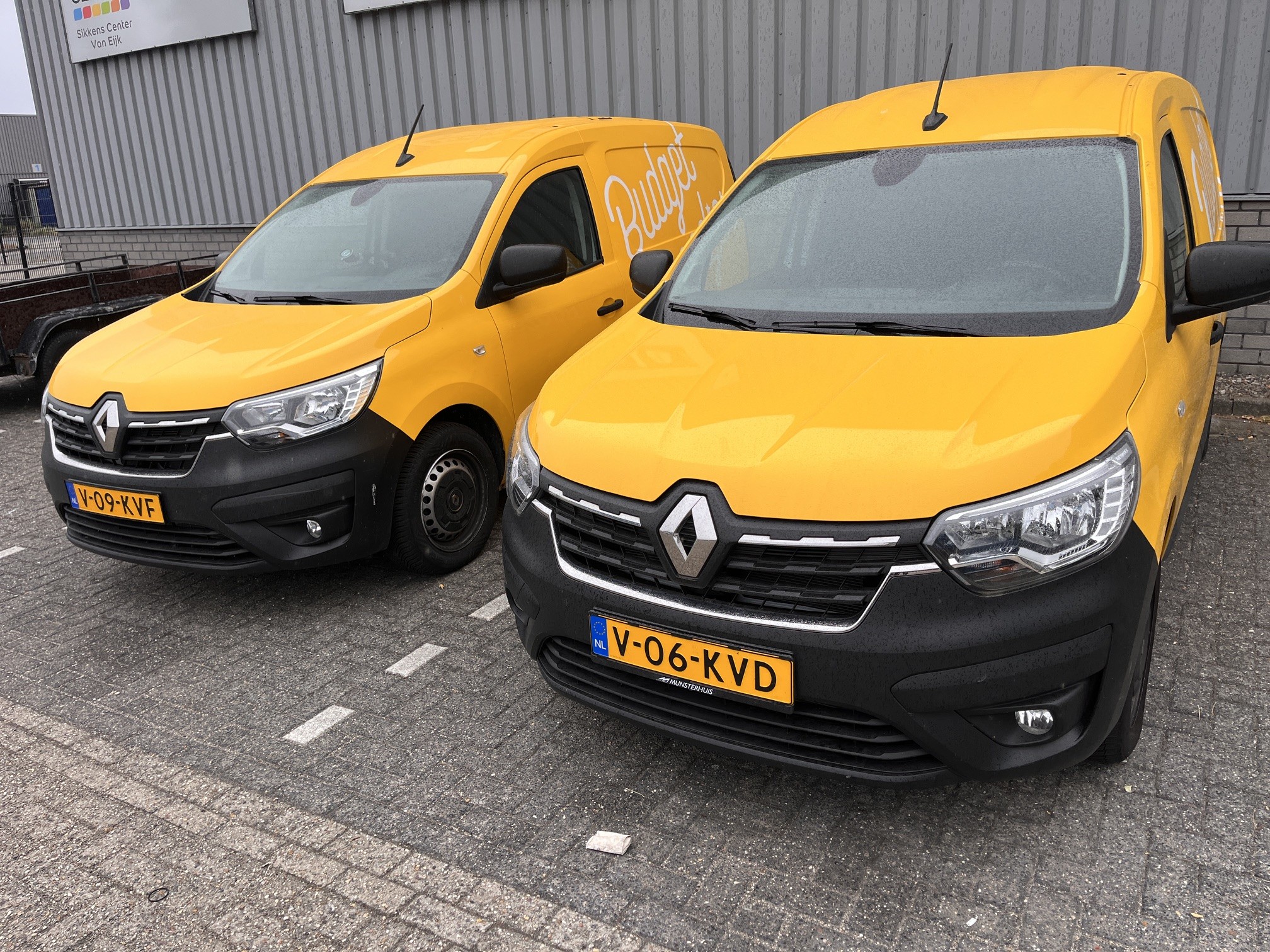 Twee gele Renault bestelwagens met deels zichtbare belettering staan geparkeerd op een stenen terrein voor een grijze loods.