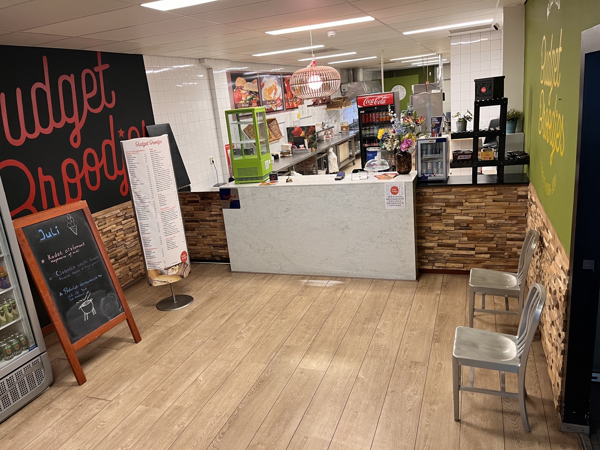 Interieur van een broodjeszaak met een witte balie, houten vloer en muren, menukaarten, metalen stoelen en zicht op de keuken.