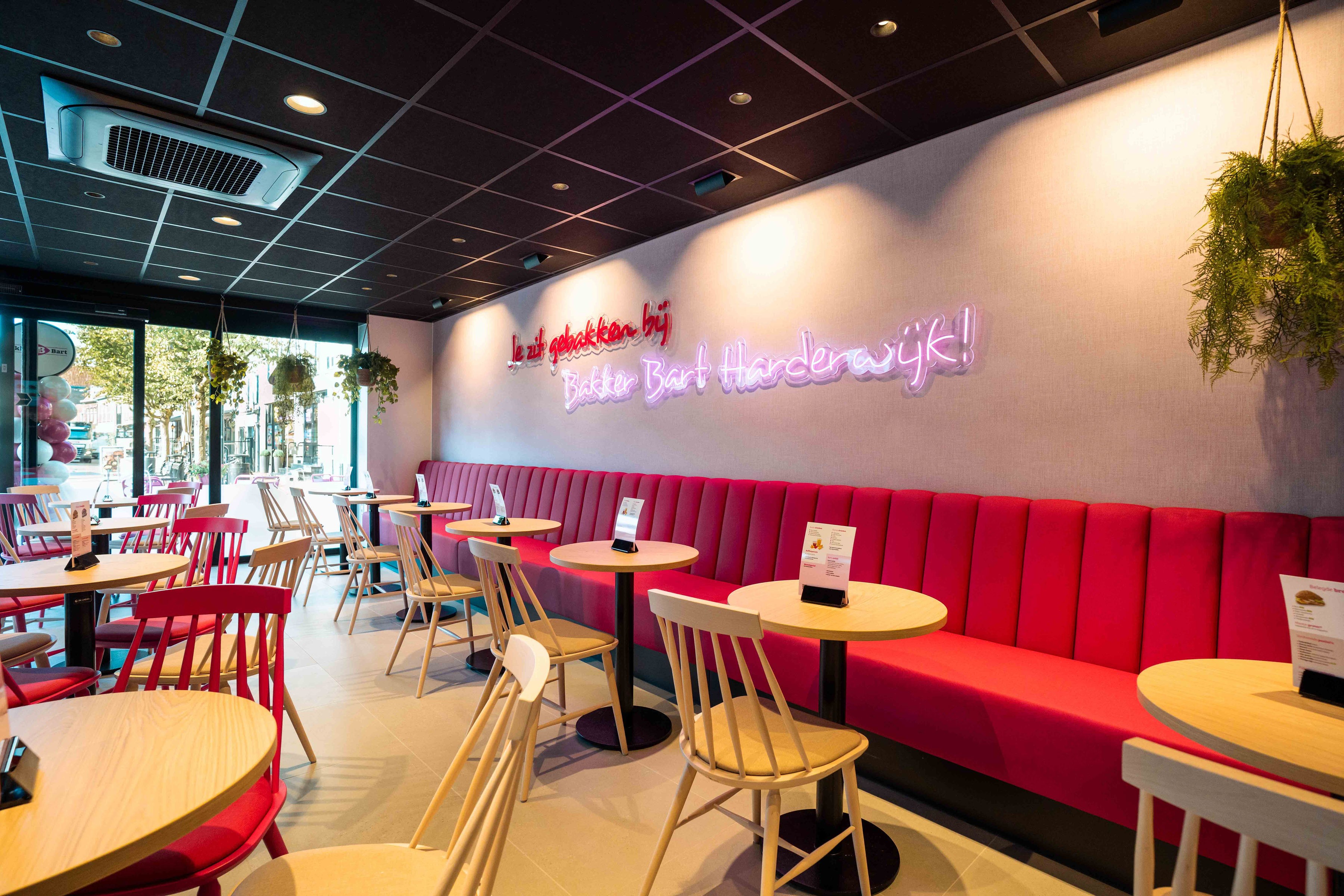 Interieur van een bakkerij met een lange roze wandbank, houten tafeltjes en een neonverlichting met de tekst Bakker Bart Harderwijk op de muur.