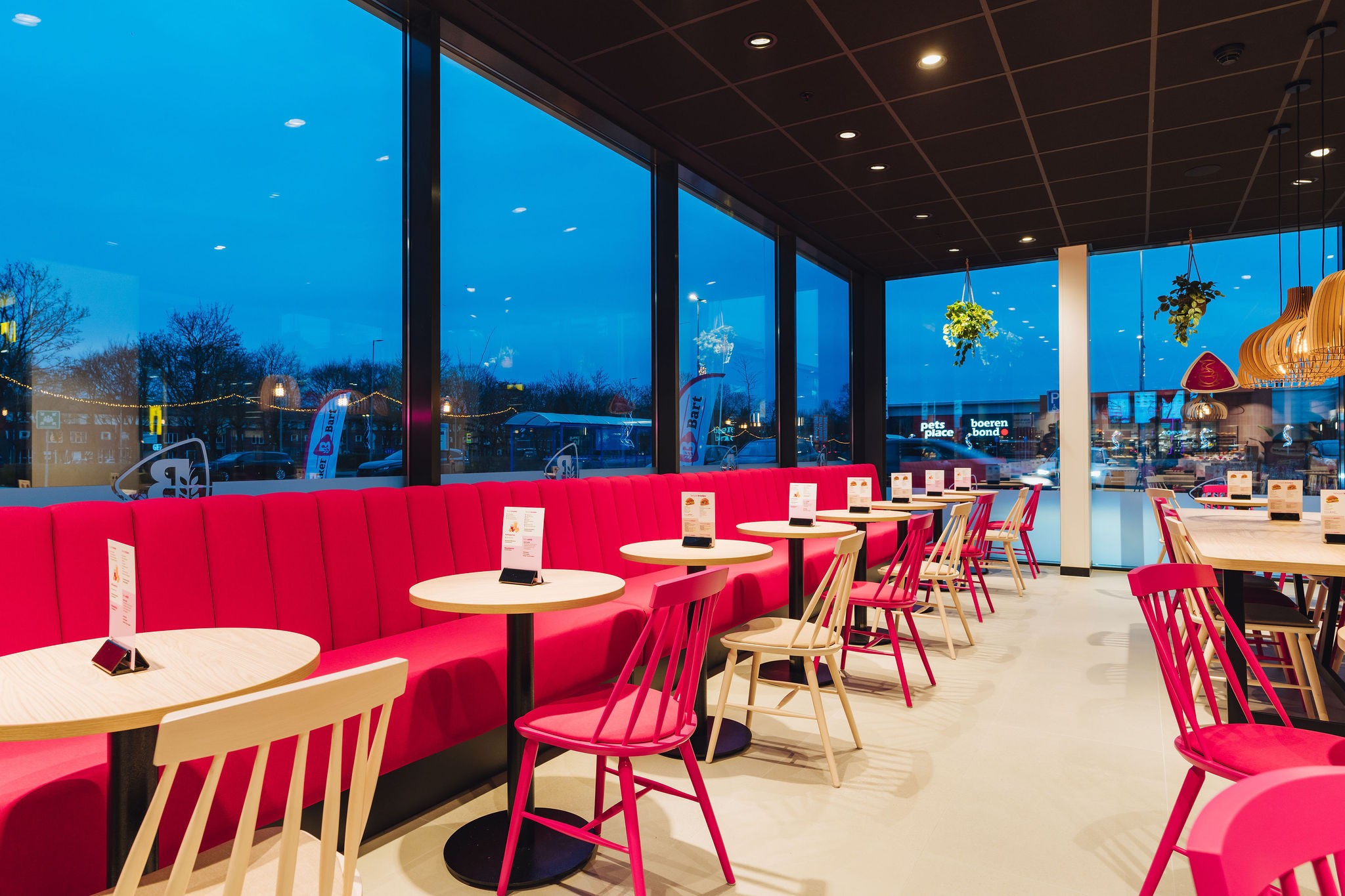 Interieur van het zitgedeelte met een lange roze bank, ronde houten tafeltjes en stoelen bij een grote raampartij.