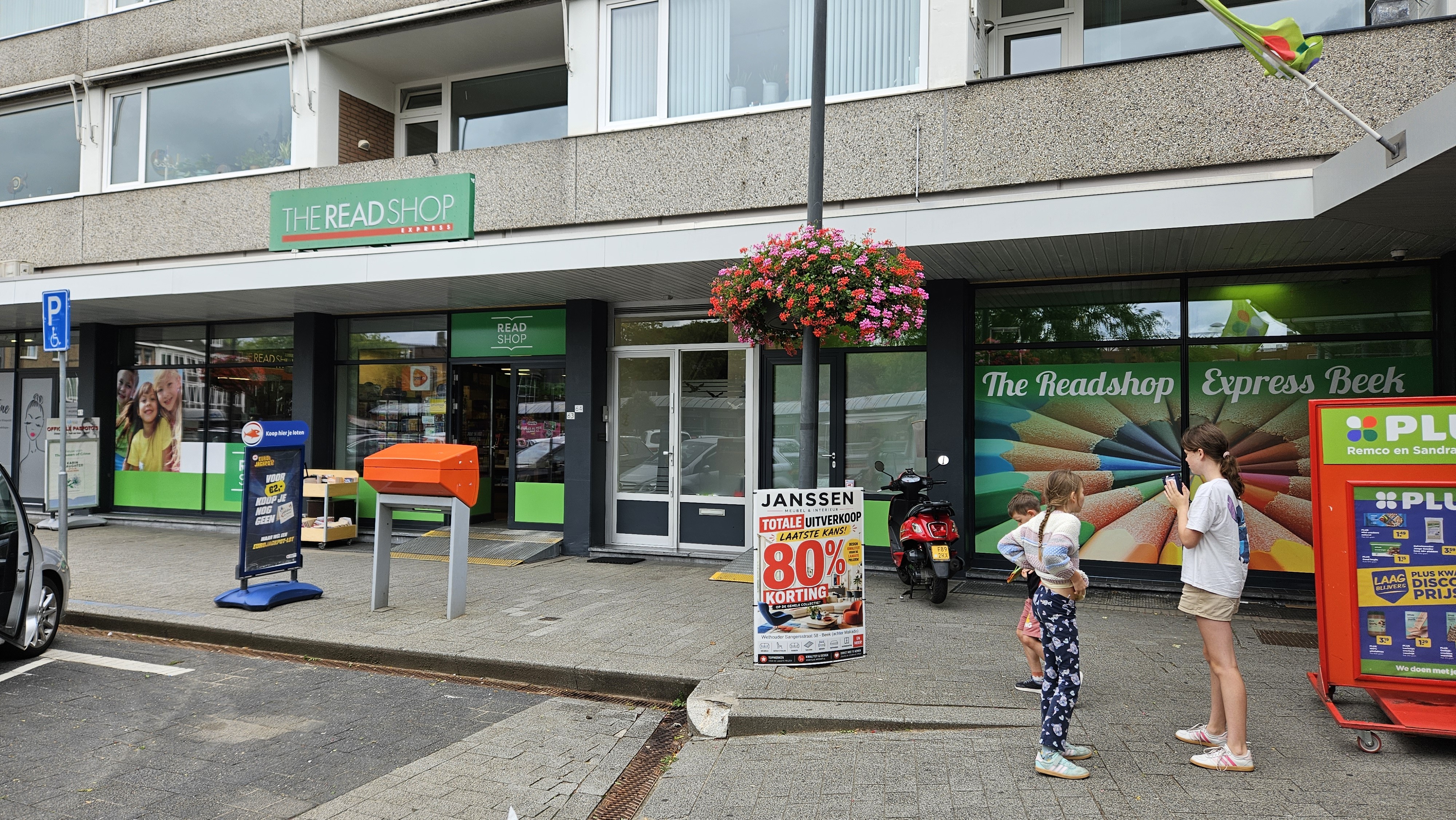 Buitenzicht van The Read Shop Express Beek met reclameborden, een rode scooter, bloembakken en kinderen op de stoep.
