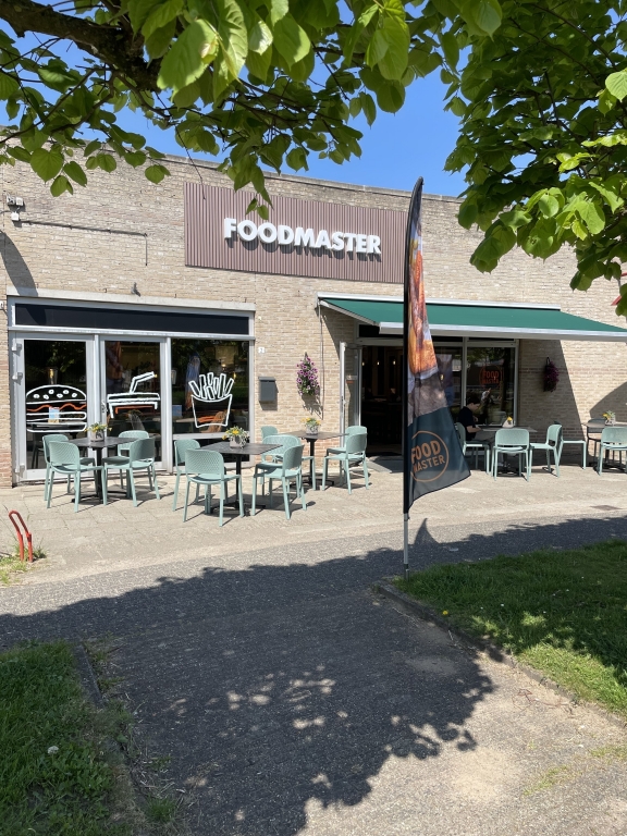 Voorgevel met de naam Foodmaster en een terras met mintgroene stoelen, tafels en een groen zonnescherm.