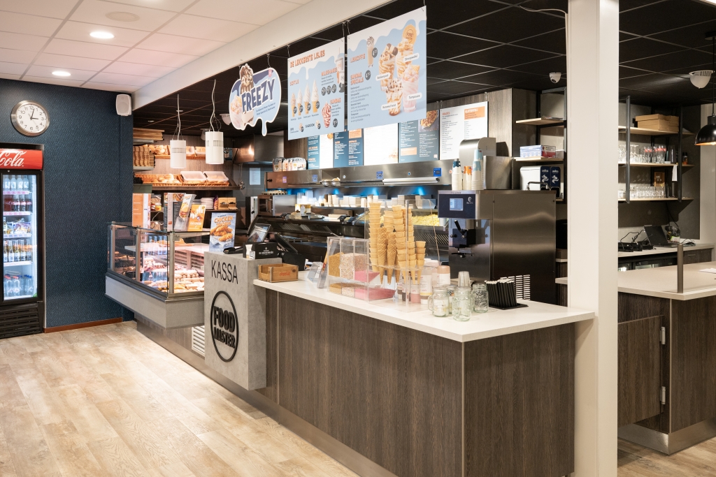 Houten counter met een kassablok voorzien van het Foodmaster-logo, een ijsmachine en menuborden aan het plafond.