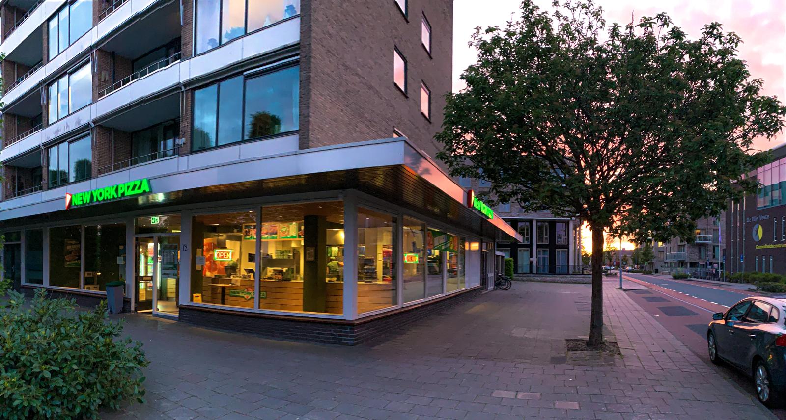 Een verlichte vestiging van New York Pizza op de begane grond van een bakstenen gebouw naast een boom en een straat bij avondschemering.