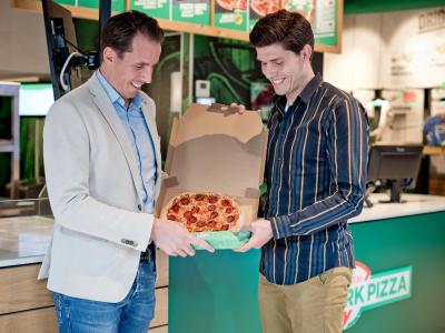 Twee mannen in een New York Pizza-vestiging bekijken samen een geopende pizzadoos met een versgebakken pizza.