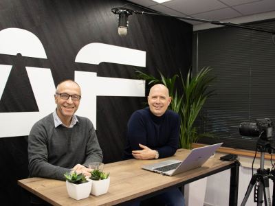 Twee mannen zitten aan een tafel met een laptop en microfoon, met het logo van Anytime Fitness op de achtergrond.