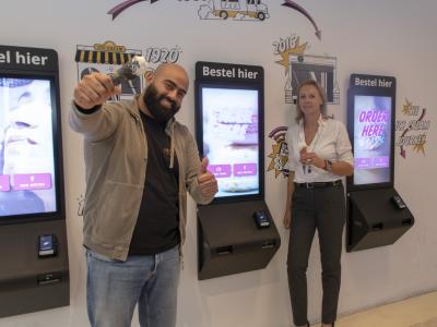 Twee mensen bij bestelzuilen in een ijssalon, waarvan één man een ijsje omhooghoudt met een duim omhoog.