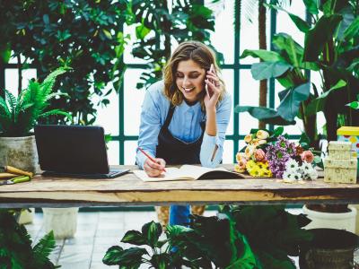 Vrouw in schort belt klantenservice terwijl ze in een bloemenwinkel notities maakt naast een laptop op een houten tafel.