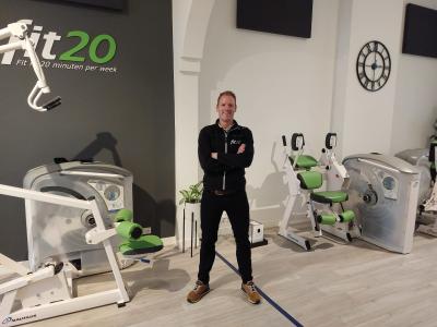 Man in fit20 studio met fitnessapparatuur, gekleed in zwarte outfit, poseert voor een grijze muur met fit20-logo.