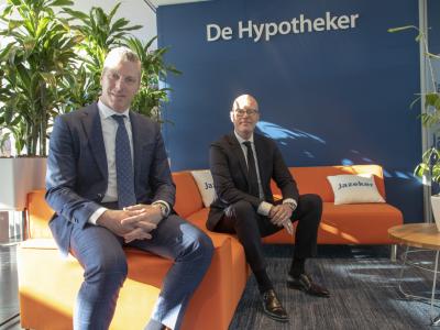 Twee mannen in pakken zitten op een oranje bank in een moderne kantoorruimte met planten en het logo van De Hypotheker.