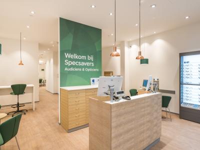 Interieur van een Specsavers-winkel met balie, groene stoelen en een rek met brillen.