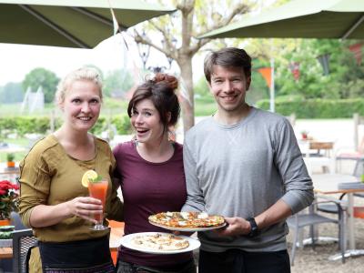Drie mensen poseren buiten met drankjes en bordjes pannenkoeken, omringd door groene natuur en decoraties.