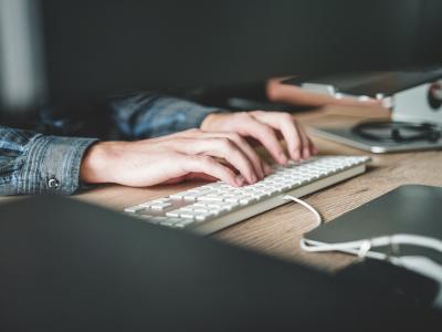 Handen typen op een wit toetsenbord naast een computer, werkend aan data-analyse op een houten bureau.