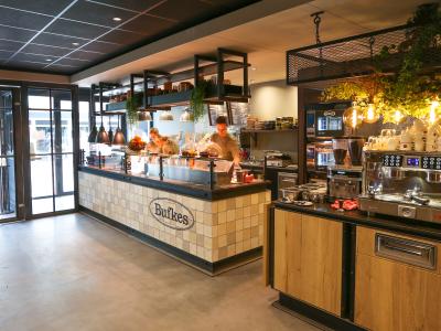 Interieur van een Bufkes-filiaal met toonbank vol gerechten, koffiezetapparaat en gezellig verlichte inrichting.