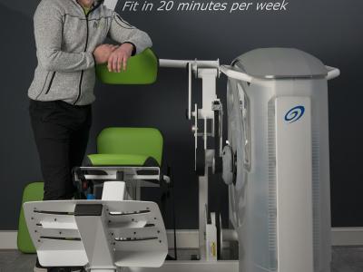 Almer Scholten staat glimlachend naast fitnessapparatuur in een Fit20 studio met het logo op de achtergrond.