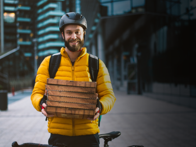 Bezorger met helm en gele jas staat naast fiets en houdt een stapel pizzadozen vast in een stedelijke omgeving.