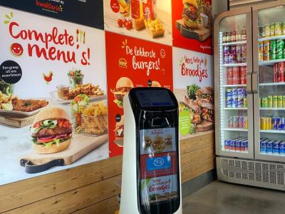 Robot Ketty in Kwalitaria Ommen naast kleurrijke reclameposters en een koeling met frisdranken.