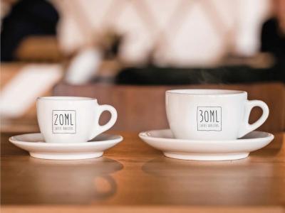 Twee witte koffiekopjes met de logo's 20ML en 30ML op een houten tafel in een caféomgeving.