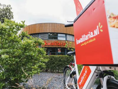 Vooraanzicht van een Kwalitaria-filiaal met een bestelfiets prominent op de voorgrond.