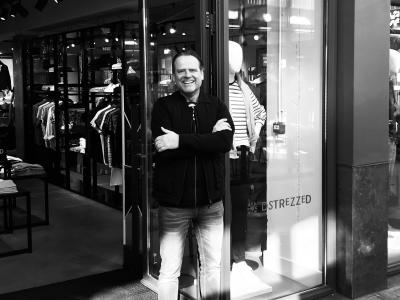 Man poseert glimlachend voor de ingang van een Dstrezzed-winkel, met kledingrekken zichtbaar binnen.