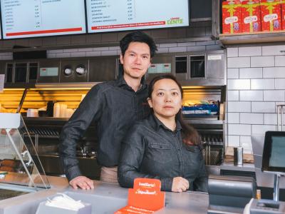 Twee mensen in uniform staan achter de toonbank in een Kwalitaria-filiaal met menuborden op de achtergrond.