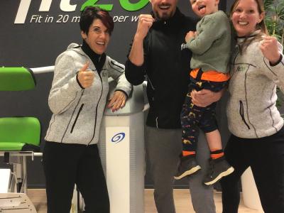 Vier mensen, waaronder een kind, poseren lachend en enthousiast bij een fit20-muur in een fitnessruimte.