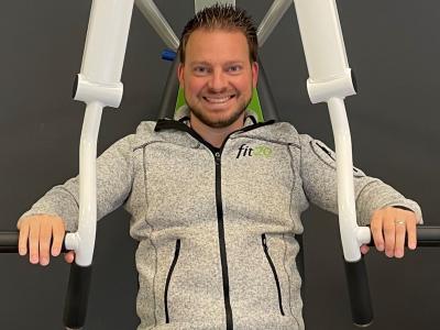 Man in grijze fit20-jas zittend op een fitnessapparaat met zwarte achtergrond.