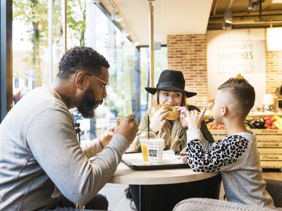 Drie mensen genieten van eten en drinken bij een tafel in een moderne foodservice-omgeving met een glazen wand.