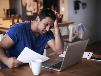 Man in blauwe trui kijkt gestrest naar laptop, met papieren in hand aan tafel met koffiekop.