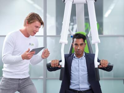 Twee mannen bij Fit20, een in trainingskleding met tablet, de ander in pak bezig op fitnessapparaat.
