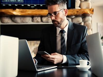 Man in pak werkt geconcentreerd met een laptop en smartphone in een moderne kantooromgeving.