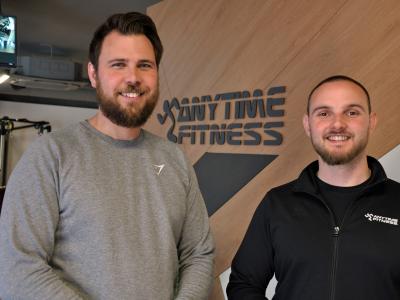 Twee mannen in sportkleding poseren voor het logo van Anytime Fitness in een moderne fitnessruimte.