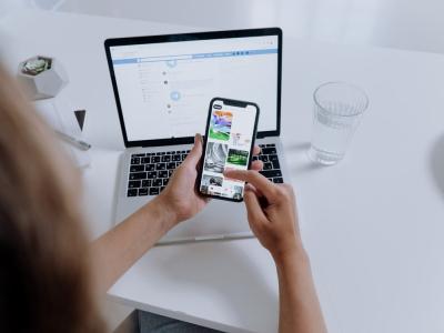 Persoon bladert door afbeeldingen op een smartphone terwijl een laptop op tafel openstaat.