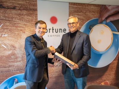 Twee mannen schudden handen voor een Fortune Coffee-banner terwijl één van hen een doos vasthoudt.