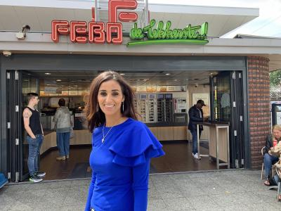Vrouw in blauwe trui glimlacht voor een FEBO-filiaal met klanten en automatiek op de achtergrond.