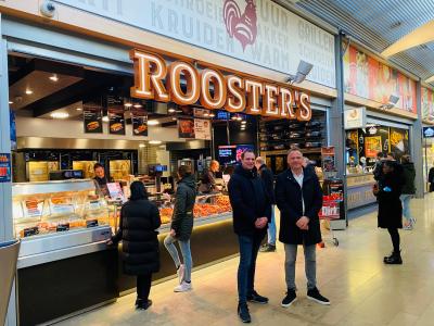 Twee mannen poseren voor een Rooster's gevogelte winkel in een overdekt winkelcentrum met klanten aan de toonbank.