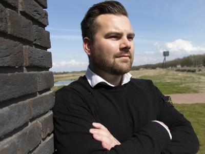 Man leunt met gekruiste armen tegen een bakstenen muur, met een landschap op de achtergrond.