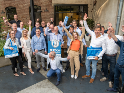 Een groep enthousiaste makelaars met verkoopborden poseert juichend voor een bakstenen gebouw.