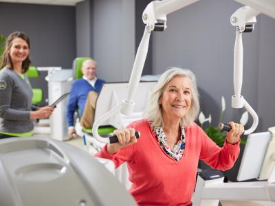 Vrouw traint op fitnesstoestel met begeleiding, met man op de achtergrond in een lichte trainingsruimte.