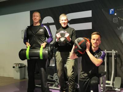 Drie mannen van verschillende leeftijden poseren met fitnessapparatuur in een sportschoolomgeving.