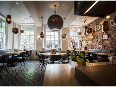 Een sfeervol ingericht restaurantinterieur met houten tafels, moderne verlichting en grote ramen met uitzicht op de straat.