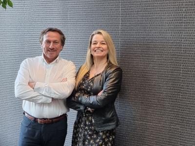 Twee mensen poseren met gekruiste armen voor een muur met een patroon in een lichte, moderne ruimte.