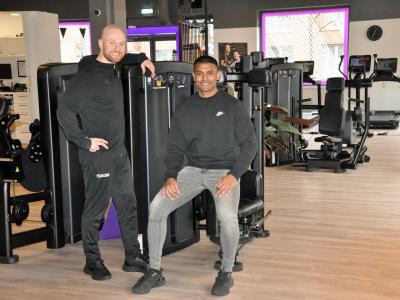 Twee mannen poseren bij fitnessapparatuur in een moderne sportschool met cardioapparaten op de achtergrond.
