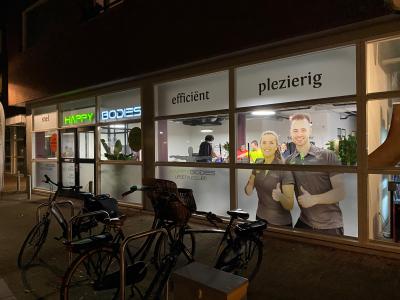 Buitenkant van sportschool Happy Bodies met fietsen en afbeeldingen van lachende mensen in het raam zichtbaar.