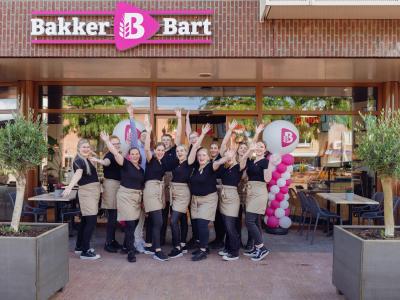 Medewerkers van Bakker Bart staan vrolijk voor de winkel met ballonnen en een bord met het logo op de achtergrond.